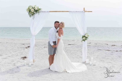 bride and groom standing in front of bamboo arbor with white draping and flowers