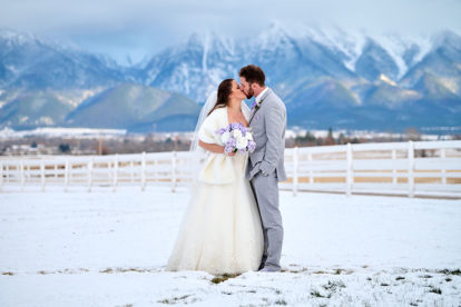 bride and groom standing in snow and kissing with mountains in the background