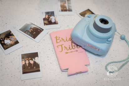 Polaroid camera with several prints and a pink koozie that says Bride Tribe