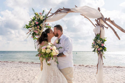 bride and groom kissing on the beach under an arbor draped with fabric and flowers