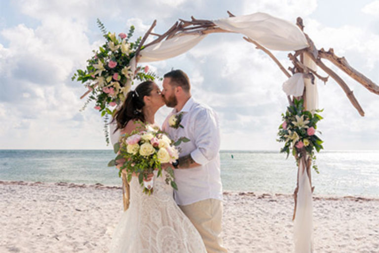 bride and groom kissing on the beach under an arbor draped with fabric and flowers