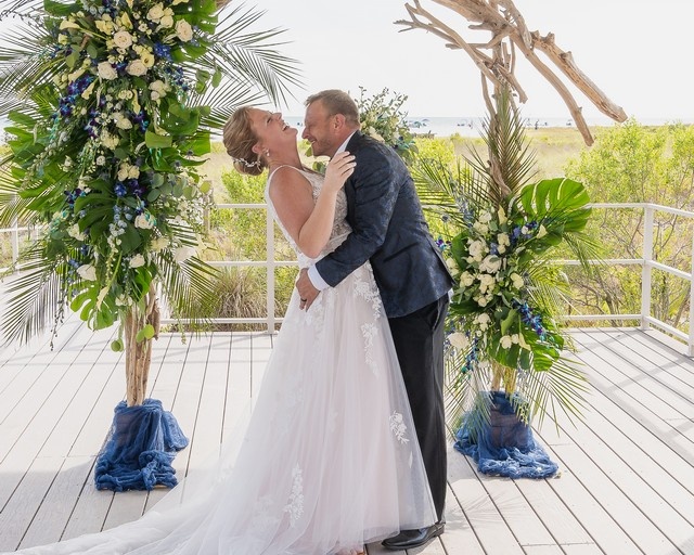 bride and groom laughing, standing under a flower-decorated arbor near the beach