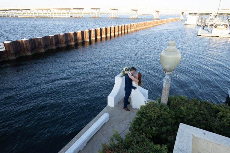 romantic aerial view of a bride and groom outdoors standing on an outcropping near the ocean with wooden pilings in the water
