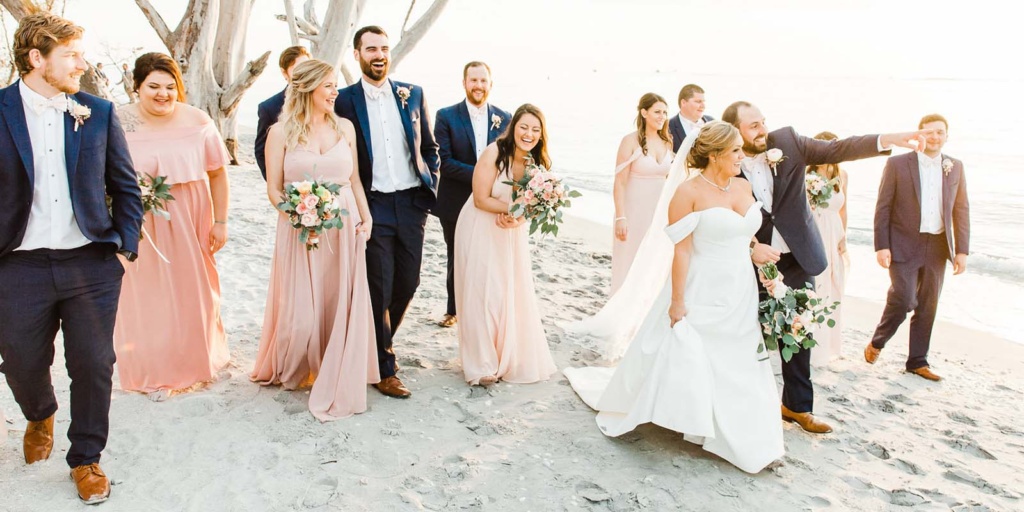 bride and groom walking, pointing, and laughing with their wedding party on the beach